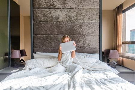 Happy young woman rests in bed and smiles at the camera while playing with laptopの写真素材