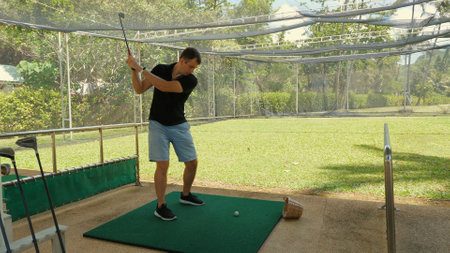Focused man practicing golf swing on driving range with greeneryの写真素材