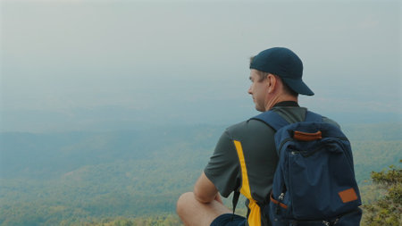 Young man with backpack sitting on mountain top, enjoying serene viewの写真素材