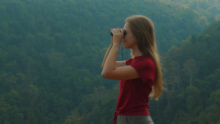 Young woman in red shirt using binoculars to observe lush, greenの写真素材