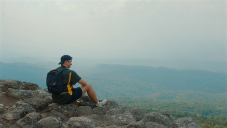 Man Sitting on Rocky Cliff Edge, Gazing at Hazy Mountain View Below,の写真素材