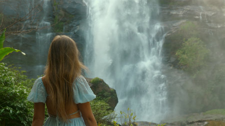 Woman in blue dress admiring majestic waterfall in lush forest,