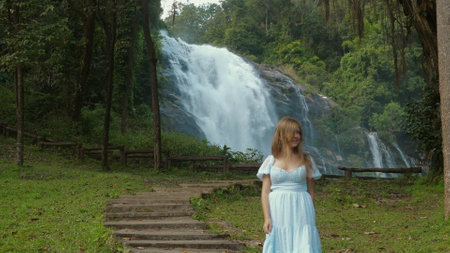 Woman in blue dress standing near waterfall in lush green forest,の写真素材