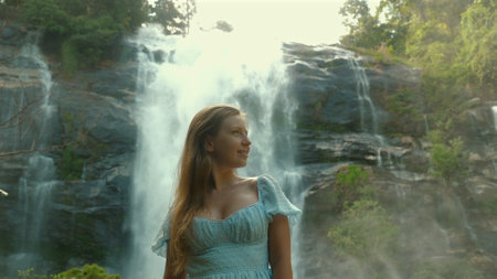 Young woman in blue dress standing in front of majestic waterfall inの写真素材