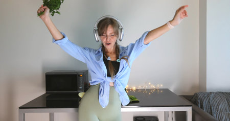 Woman dancing in kitchen with headphones, holding fresh greens, joyfulの写真素材