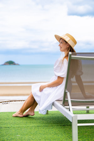Woman sitting on deck chair by ocean in straw hat and white dressの写真素材