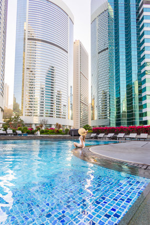 Woman in swimsuit sits by rooftop pool in modern cityscape.