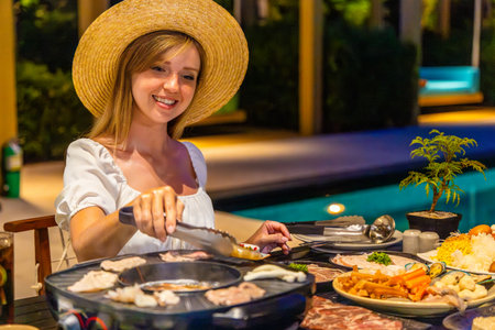 Woman at outdoor barbecue, smiling in hat, grilling food near poolsideの写真素材