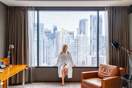 Woman in hotel room sits by large window overlooking urban skyline.の写真素材