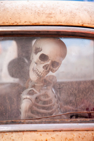 Close up photo of a human skeleton model sitting inside an old rusted car seen through a dirty window.. The scene has an eerie surreal and slightly humorous mood suitable for themes related to halloween spooky concepts abandoned places and dark creative photographyの写真素材