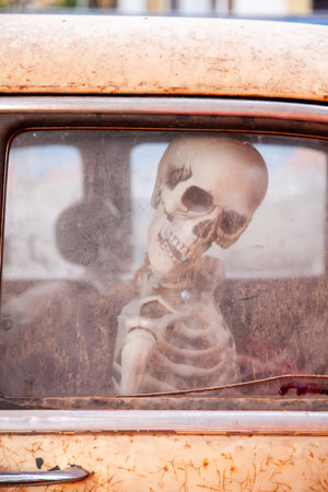 Close up photo of a human skeleton model sitting inside an old rusted car seen through a dirty window.. The scene has an eerie surreal and slightly humorous mood suitable for themes related to halloween spooky concepts abandoned places and dark creative photographyの写真素材
