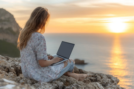 Young woman sitting on a rock by the sea using laptop at sunsetの素材
