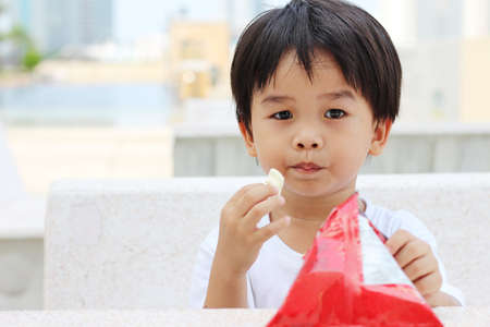 Asian boy sitting on a concrete chair and eating a snack.の写真素材