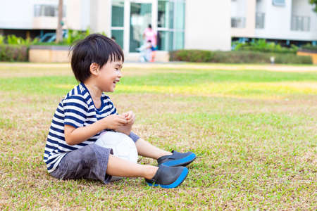 Asian little boy sitting alone with a soccer ball on the lawn in the public park.の写真素材