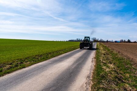 road along which goes an old tractor on wood with green fields around and blue skyの写真素材