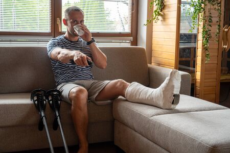 Young man watching TV in bandage on sofa and drinks coffeeの写真素材