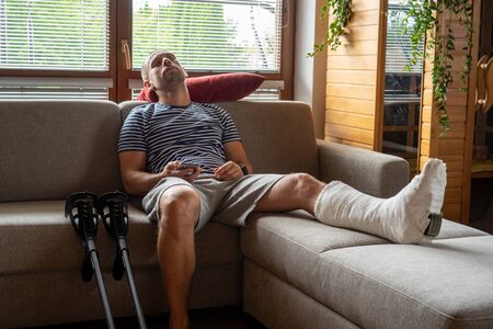 Side view of a young handsome bearded man in casual clothes sleeps on the couch in a room at home. with feet in plasterの写真素材