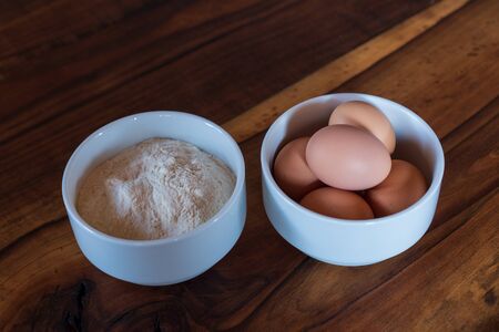 eggs and flour in small white bowls on a wooden backgroundの写真素材