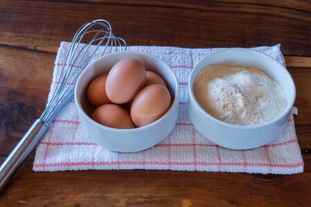 Raw chicken eggs on white bowl with flour and with whisk on wooden background. Ready to start the cooking conceptの写真素材