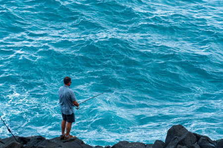 Active recreation in nature. A man with a fishing rod is fishing on the rocks near the sea. A fisherman catches fish at sunset.の写真素材