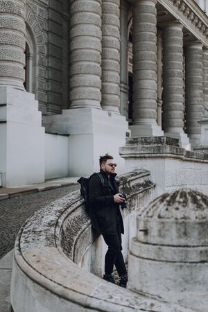 Stylish guy with black backpack standing outdoors and using camera with building on background. Travel conceptの写真素材