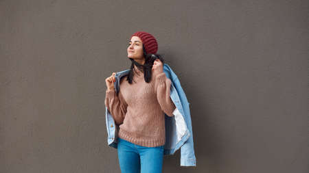 Pleasant young lady in stylish attire wrapping herself up in denim coat while looking aside near a...
