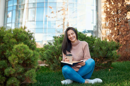 Nice-looking female student laughing enjoying funny book on a cozy green lawn nearby a tall city towerの写真素材