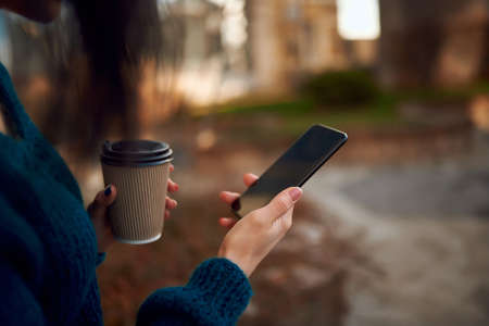Cropped shot of young woman hands with a smartphone and disposable cup of hot drink on defocused...
