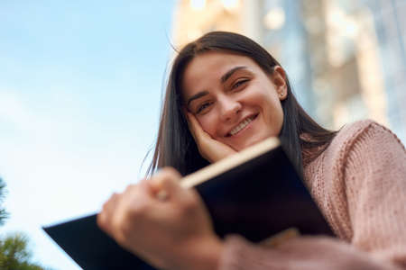 Beautiful young female looking over the pages of book and holding in hand with smile on blurred...