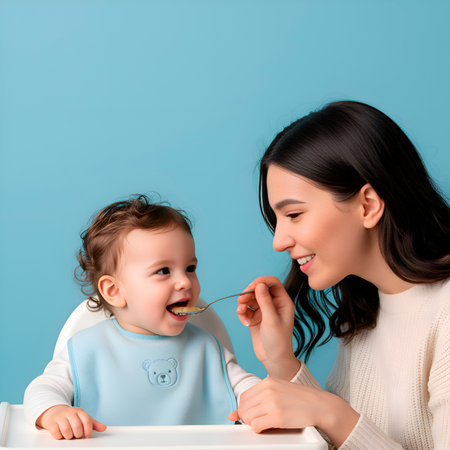 A loving mother smiles while spoon-feeding her happy baby in a high chair, illustrating bonding, nutrition, and early childhood development against a blue studio background.の素材