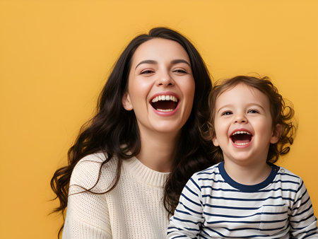 Portrait of a beautiful mother and her excited young child genuinely laughing together against a vibrant yellow studio background showing pure joy.の素材