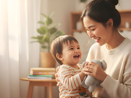 Happy Asian mother and baby share a joyful, intimate moment playing with a plush toy in the bright, sunlit living room.の素材