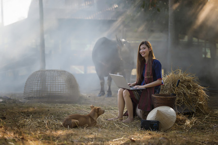 Young Woman Farmer Sitting And Play Notebook And Dog Looking Her With Buffalo Backgroundの写真素材