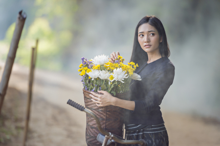 Beautiful Asian woman in local traditional dress with old bicycle and flower basket on the green summer field.の写真素材