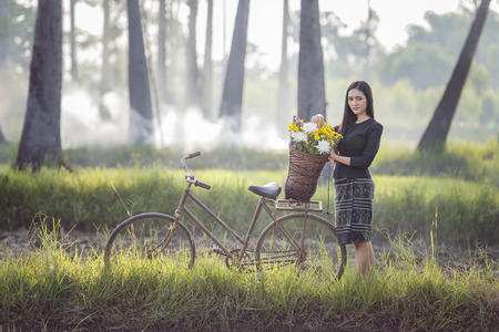 Beautiful Asian woman in local traditional dress with old bicycle and flower basket on the green summer field.の写真素材