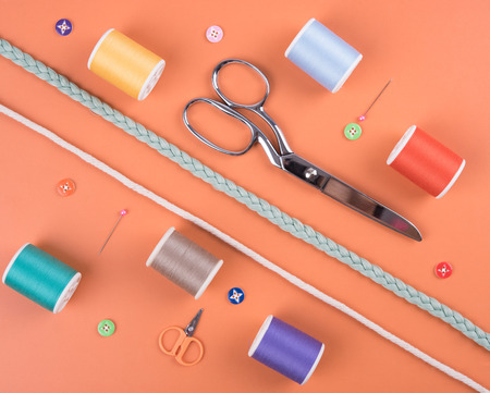 closeup needles, thread spool, scissors, button on orange wooden background.