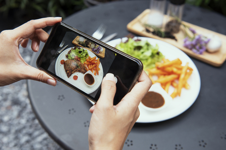 blogger hands with smartphone photographing pork chop steak with vegetable salad and potato chips on white plate in the garden. top view.の写真素材