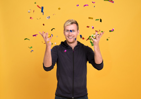 Portrait of a cheerful man standing standing under confetti rain and celebrating isolated over yellow background.の写真素材