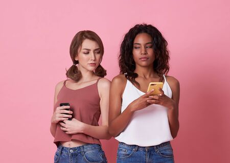 Image of curious woman spying and peeking at smartphone of her friend isolated over pink background.