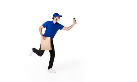 Asian delivery man wearing face mask in blue uniform running with paper bag and takeaway coffee isolated over white background. express delivery service during covid19.の写真素材
