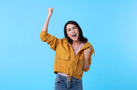 Cheerful young Asian woman raising his fists with smiling delighted face, yes gesture, celebrating...