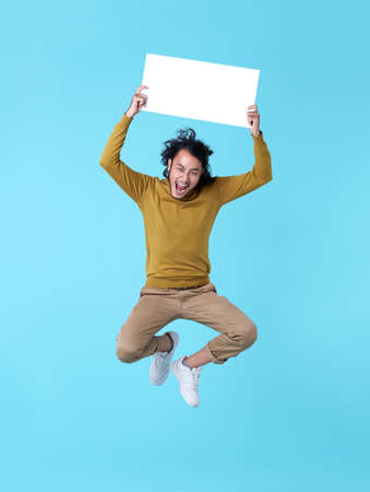 Happy energetic young Asian man jumping in mid-air holding blank speech bubbles on blue background.