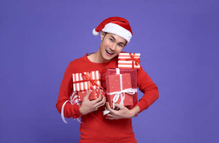 Young asian man in red casual attire wearing Santa hat and holding stack of presents with smile face on purple background.Happy new year concept.の写真素材