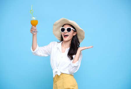 Happy Asian woman in summer casual clothes wearing straw hat, sunglasses holding glass of fresh fruit juice drink isolated on bright blue backgroud.の写真素材