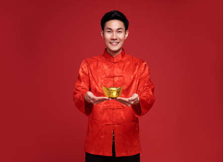 Happy Chinese new year. Asian man wearing in traditional costume holding gold ingot isolated on red background.の写真素材