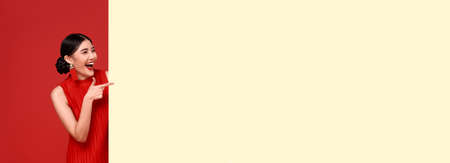 Happy asian woman in red casual attire with hand pointing blank announcement banner in red background.の写真素材