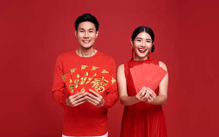Happy asian couple in red casual attire with holding angpao or red packet monetary gift of congratulation greeting happy new year 2021 on bright red background. text means great luck.の写真素材