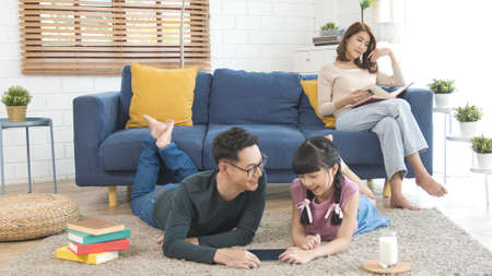 Relax Asian mother reading a book on sofa and father with daughter using tablet in living room at home.の写真素材