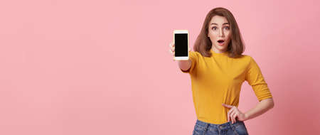 Happy young woman showing at blank screen mobile phone and hand gesture success isolated over pink...