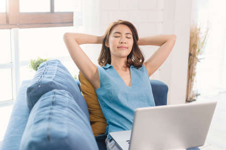 Asian businesswoman using laptop work at home stretch relax breathe fresh air on sofa. social...